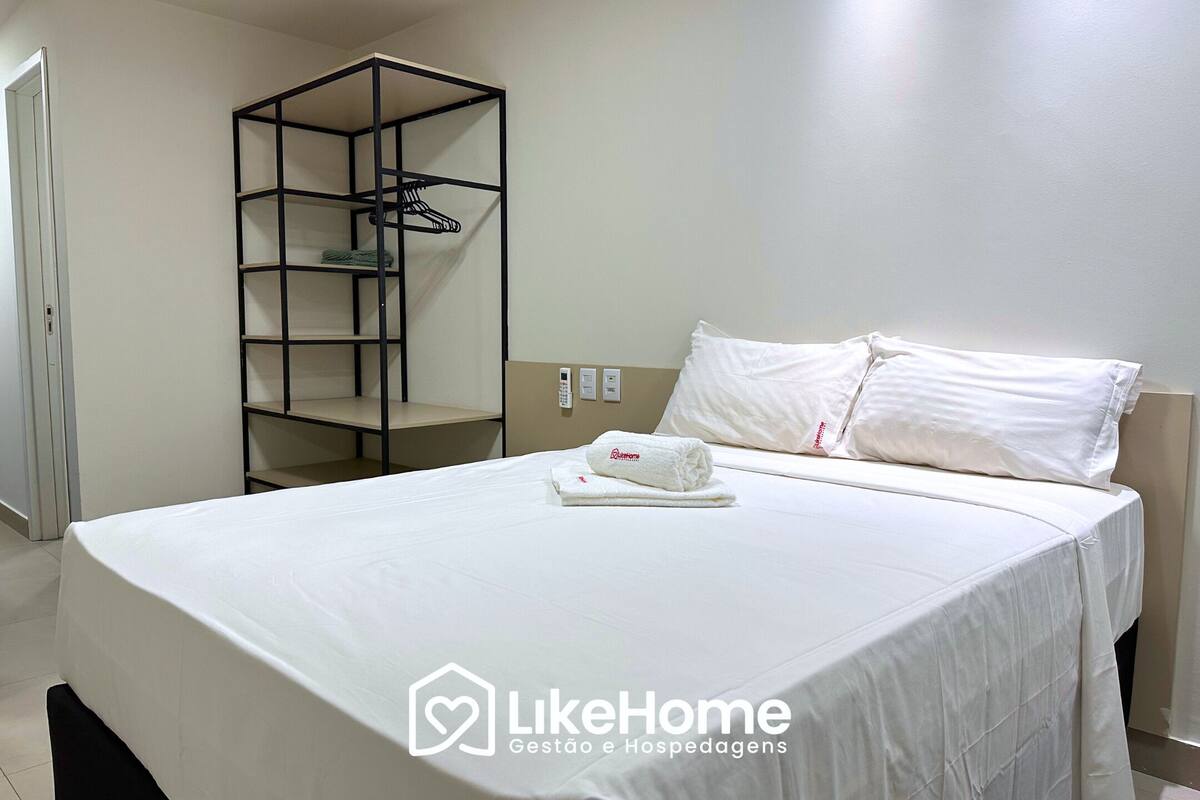 A neatly made bed with a white comforter and a folded towel, a black metal shelf unit, and a white wall with a power outlet.