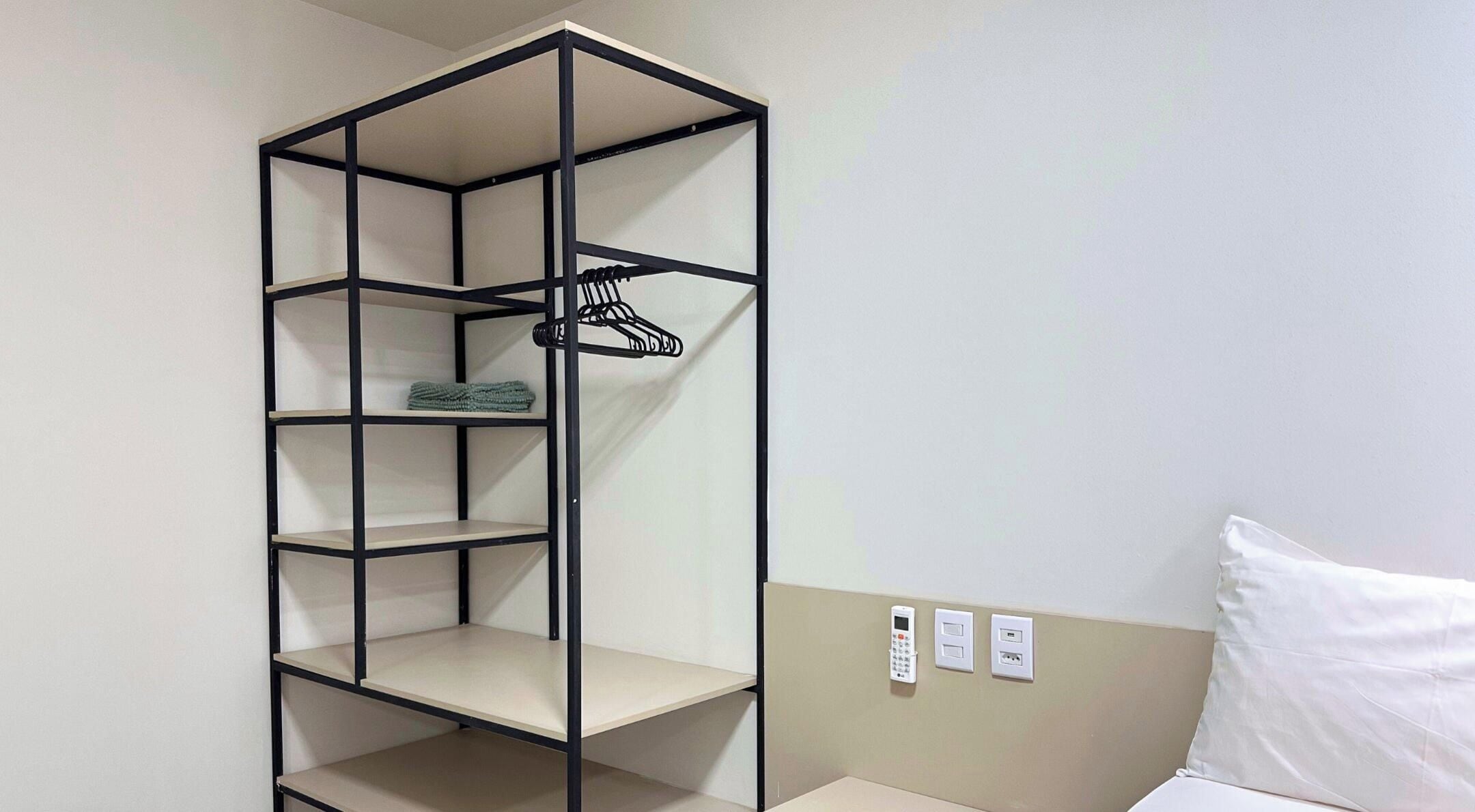 Une pièce dotée d’une armoire intégrée avec plusieurs étagères et une tringle à vêtements, d’un lit avec un oreiller blanc et d’une prise de courant murale.