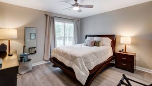 Un dormitorio con cama, mesitas de noche, ventilador de techo, ventana con cortinas y mesita auxiliar.