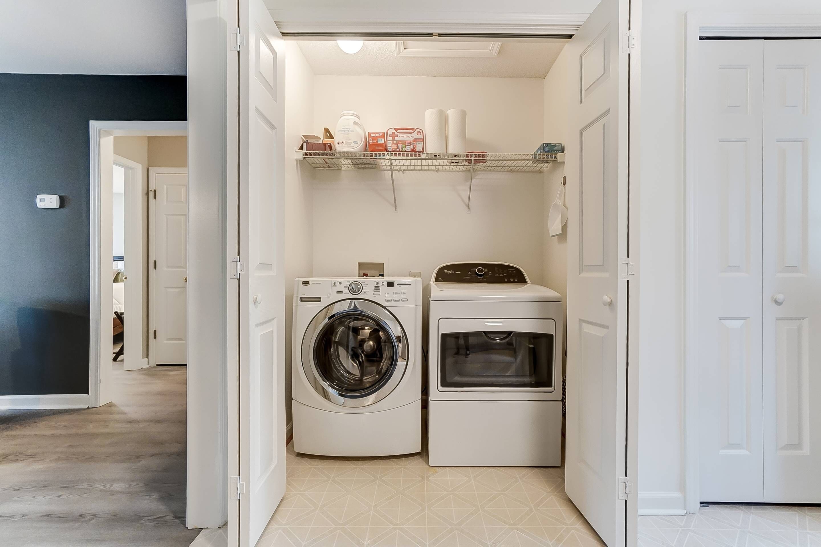 Une buanderie équipée d’un lave-linge et d’un sèche-linge à chargement frontal, d’une étagère pour les articles de toilette et d’une porte donnant sur une autre pièce.