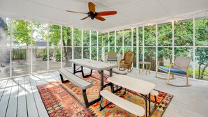 Une terrasse couverte avec une table et des bancs, entourée de grandes fenêtres et dotée d’un ventilateur de plafond.