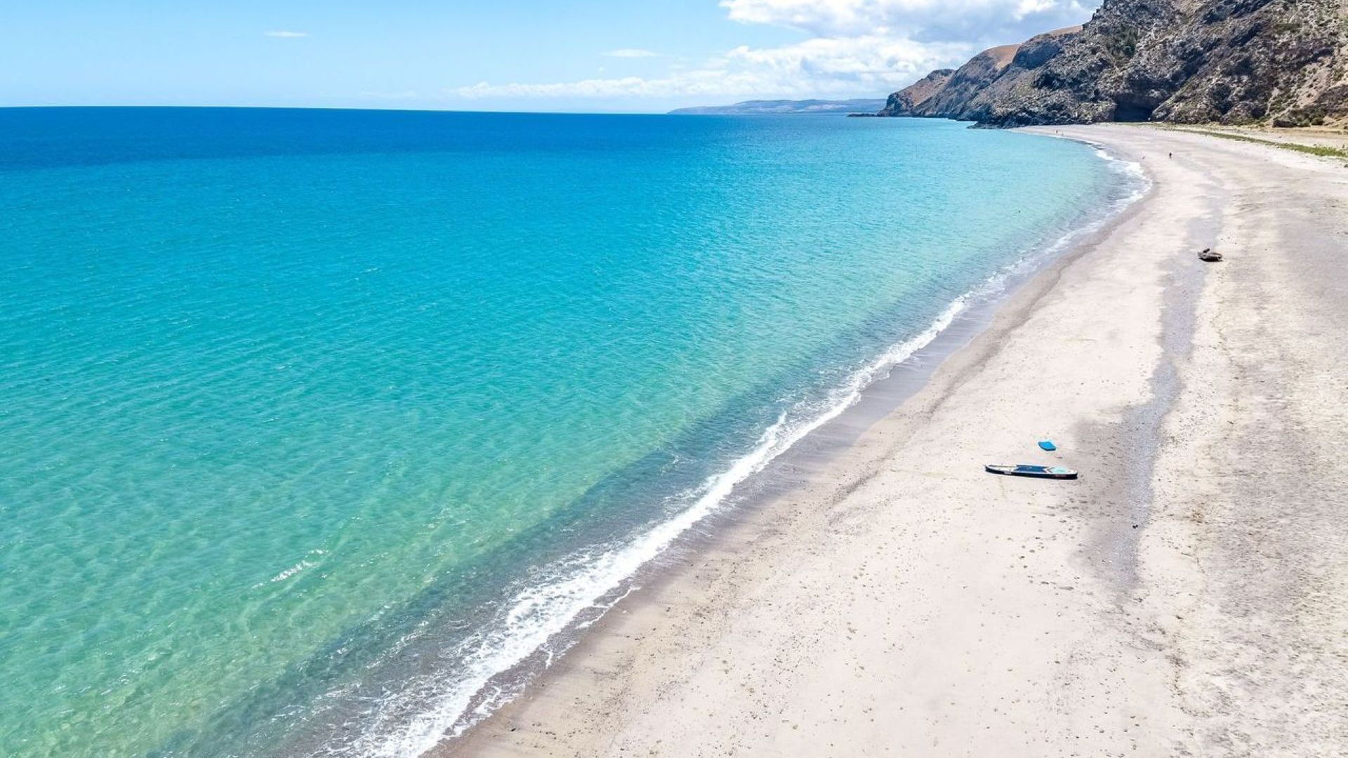 Una playa con agua turquesa cristalina, una orilla arenosa y acantilados rocosos al fondo.