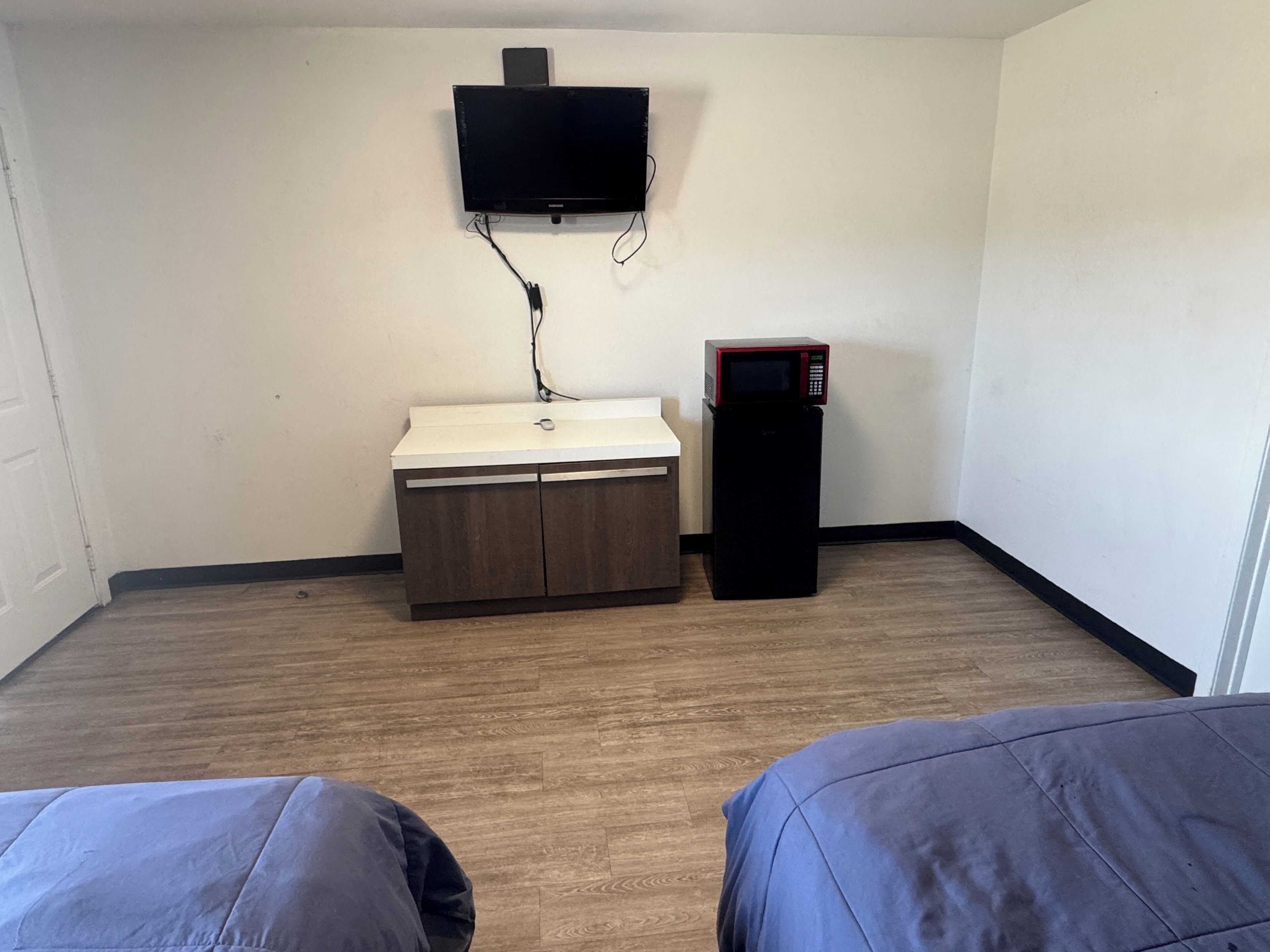 A small room with a bed, a desk with a sink, a microwave, and a TV mounted on the wall.
