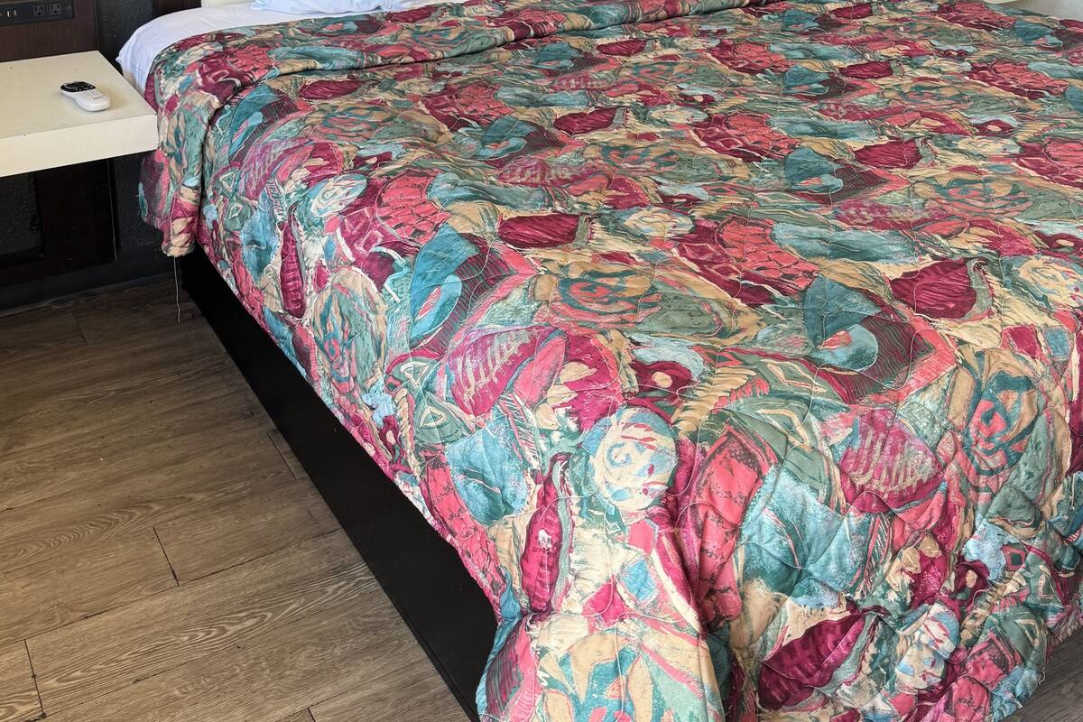 Una habitació d'hotel amb un llit gran, nínolls blancs, un llençol de llit colorit, un capçal de llit de fusta, una tauleta de nit amb un comandament a distància i un paviment de fusta.