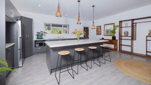 A modern kitchen with grey cabinets, a large island with bar stools, pendant lighting, and a view of greenery outside.