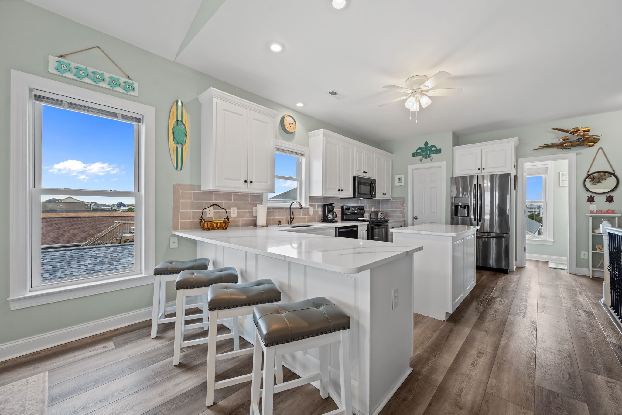 A modern kitchen with white cabinetry, a large island with stools, a view of the beach, and a ceiling fan.