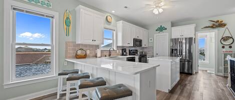 A modern kitchen with white cabinetry, a large island with stools, a view of the beach, and a ceiling fan.