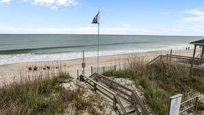 Ein Strand mit einer hölzernen Treppe, die zum Sand führt, einer Fahne an einem Mast und einem Holzzaun.
