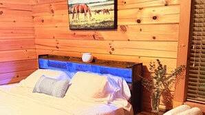 Een houten slaapkamer met een bed, een nachtkastje, een vaas met een plant en een raam met jaloezieën.