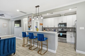 A modern kitchen with white cabinets, a stainless steel refrigerator, and a black stove. There are three blue bar stools and a white island with a vase and a coffee machine. - NRG Rest & Rally | Sleeps 12 (Houston)