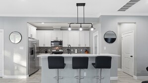 A modern kitchen with a central island, bar stools, stainless steel appliances, and a pendant light fixture. - NRG Rest & Rally | Sleeps 12 (Houston)