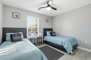A bedroom with two beds, a ceiling fan, a window with blinds, and a small table. - NRG Rest & Rally | Sleeps 12 (Houston)