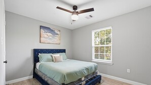A bedroom with a bed, a ceiling fan, a window with blinds, and a painting on the wall. - NRG Rest & Rally | Sleeps 12 (Houston)