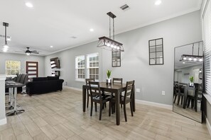 A spacious living room with a dark brown sofa, a dining area with a wooden table and chairs, and a kitchen in the background. - NRG Rest & Rally | Sleeps 12 (Houston)