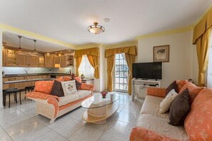 Uma sala de estar com um sofá, uma mesa de centro e uma televisão. Cozinha visível ao fundo, com armários e uma janela.