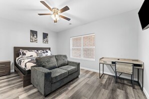 Quarto com cama, sofá cinza, escrivaninha e ventilador de teto.