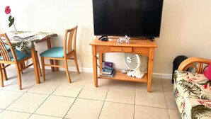 Una habitació amb escriptori de fusta, dues cadires, televisió, ventilador i un llit amb estampa floral.