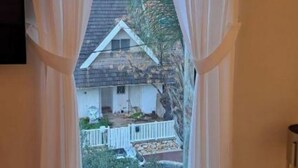 Une vue à travers une fenêtre avec des rideaux, dévoilant une maison avec une clôture blanche, un toit en pente et quelques plantes.