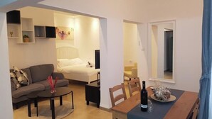 A modern living room with a sofa, coffee table, and a dining area with a table and chairs. There is a bed with a headboard in the background. - אבני החושן- Avnei Hoshen - Two-Bedroom Apartment (Nahariyya)