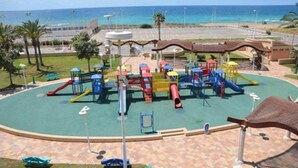 A playground with various play structures, a paved area, and a view of the ocean. - אבני החושן- Avnei Hoshen - Two-Bedroom Apartment (Nahariyya)