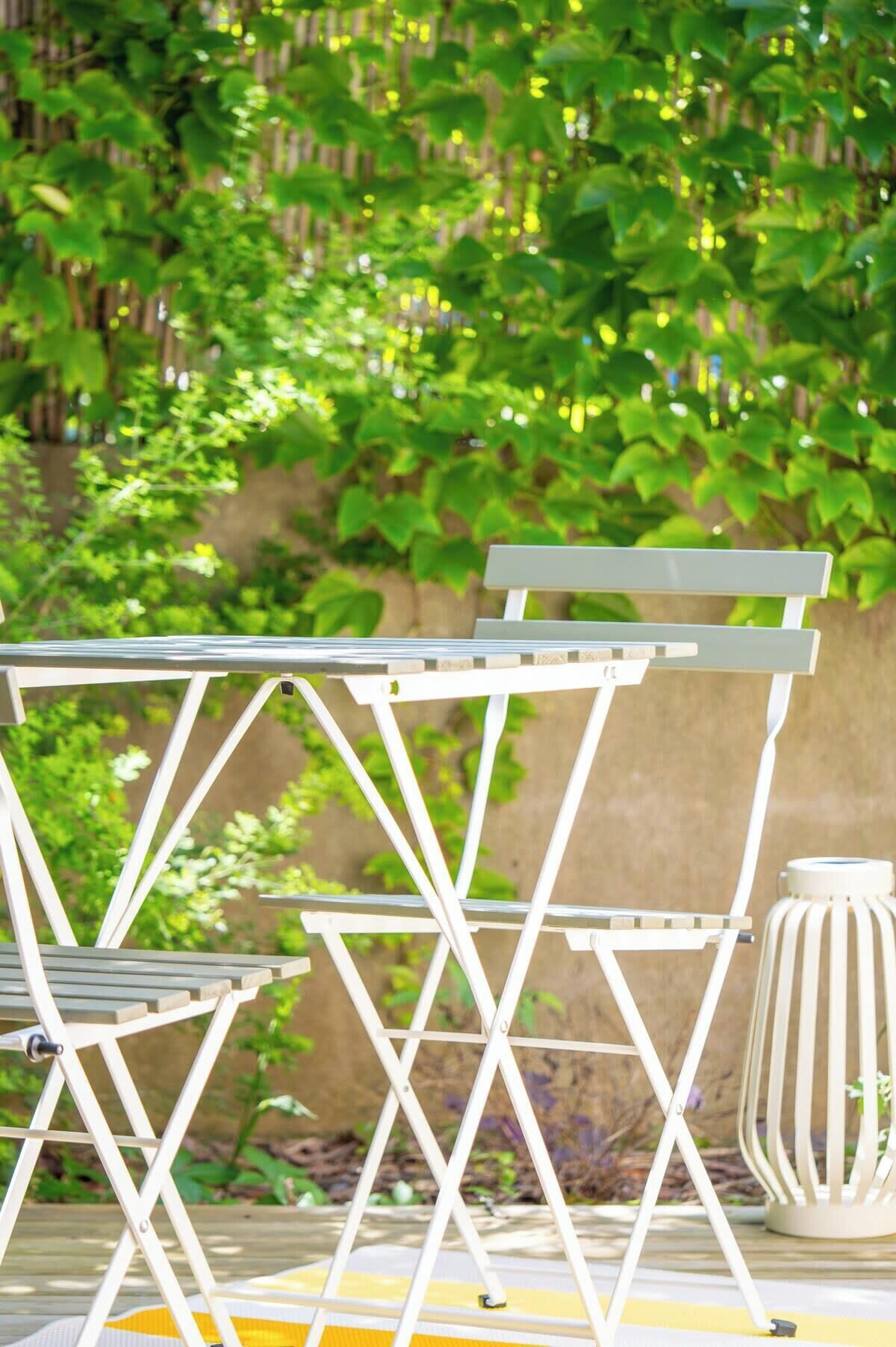 A white metal folding table and chairs set outdoors with a green leafy background.