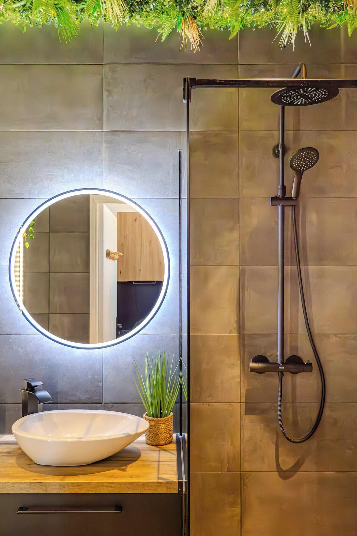Une salle de bain moderne avec un miroir rond, un lavabo sur colonne, une douchette à main et une plante en pot.