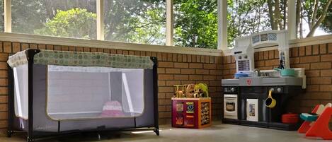A playpen with a mesh top, a toy kitchen set, and a window with a view of trees.