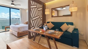 Een moderne hotelkamer met een bed, een eettafel gedekt voor twee personen, een blauwe bank en een groot raam met uitzicht op zee.