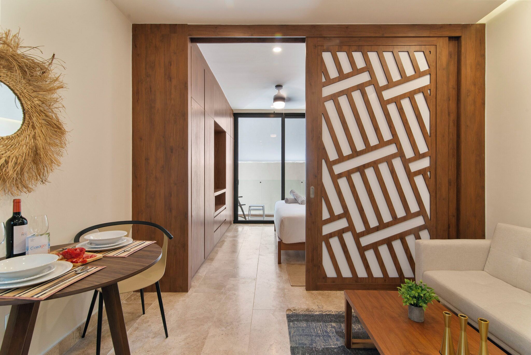 A modern living room with a sofa, a dining table set with plates and cutlery, a decorative mirror, and a wooden partition with geometric patterns.