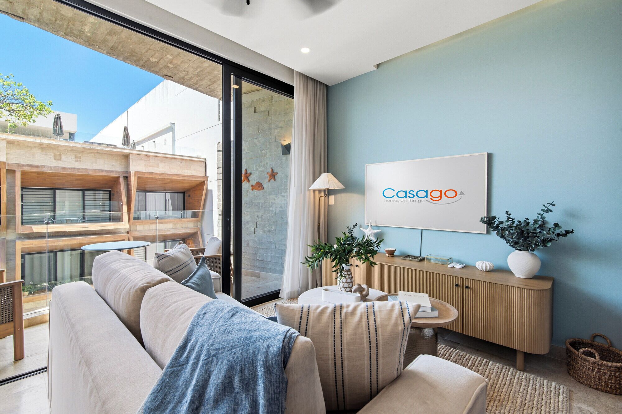 A modern living room with a sofa, a side table, a TV displaying 'Casago homes on the go,' and a view of an outdoor balcony.