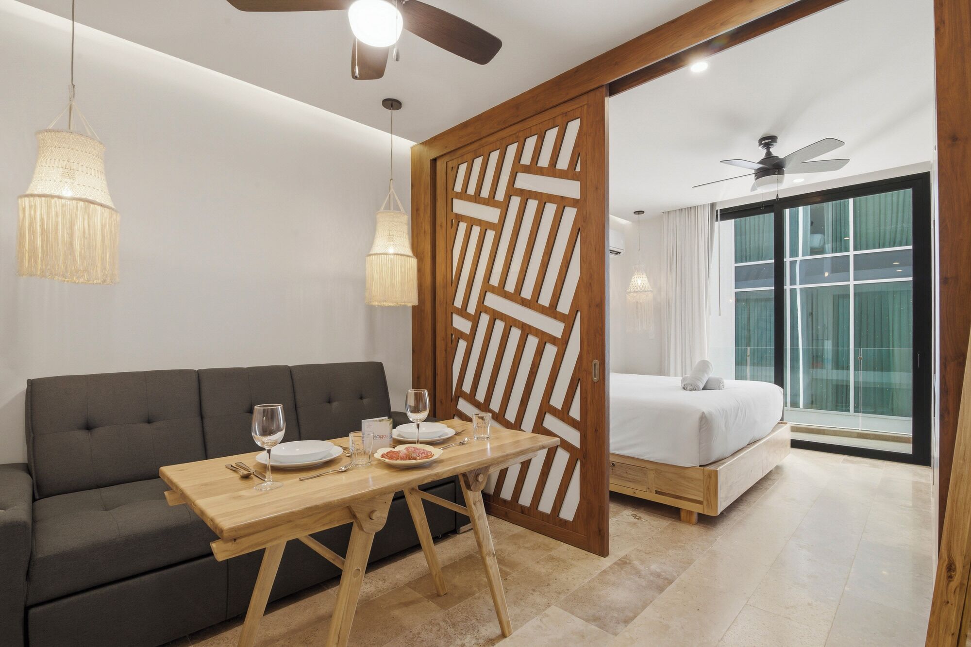 A modern hotel room with a sofa, a wooden dining table, a bed, and a large window.