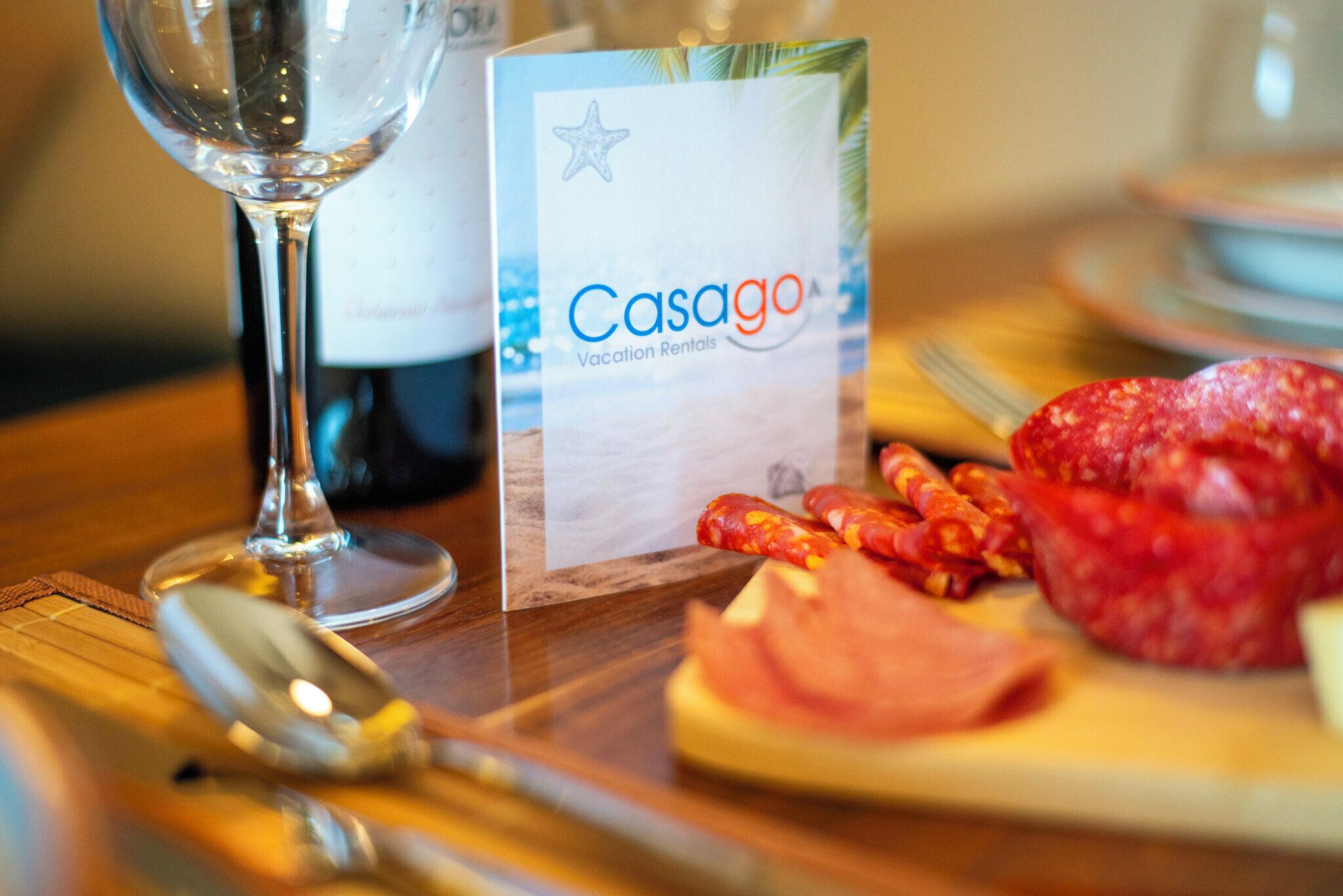 Una mesa con una copa de vino, una cuchara, un paquete con la etiqueta “Casago Vacation Rentals” y algunos alimentos.