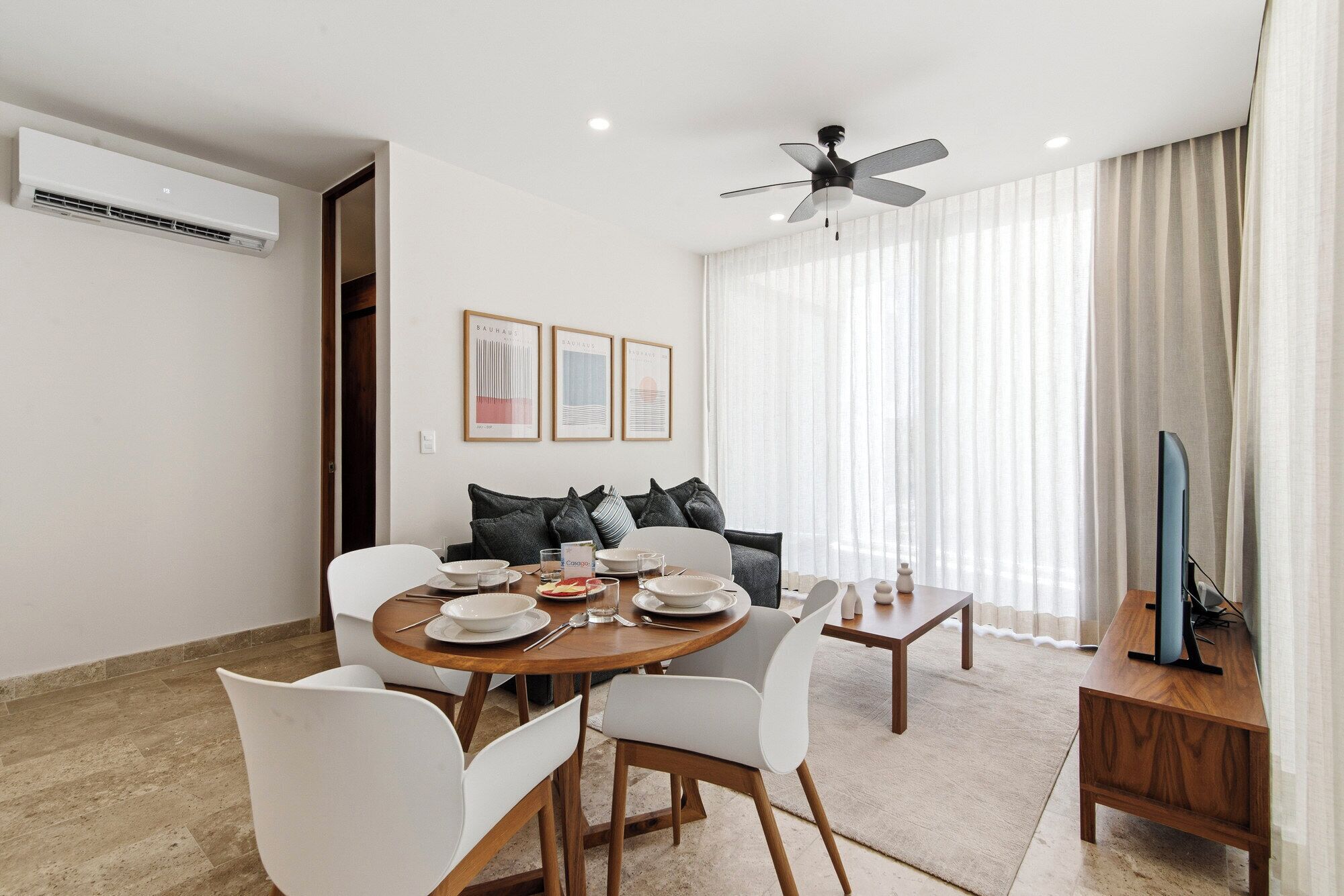 A dining area with a round table, white chairs, a sofa with cushions, a ceiling fan, and a TV on a wooden stand.