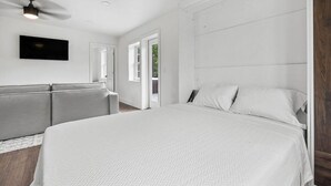 A modern bedroom with a large bed, a grey sofa, a ceiling fan, and a TV mounted on the wall. - 1 Bedroom Anchor Cottage 2 - One bedroom, one murphy bed, one sleeper sofa, with kitchenette (Allons)