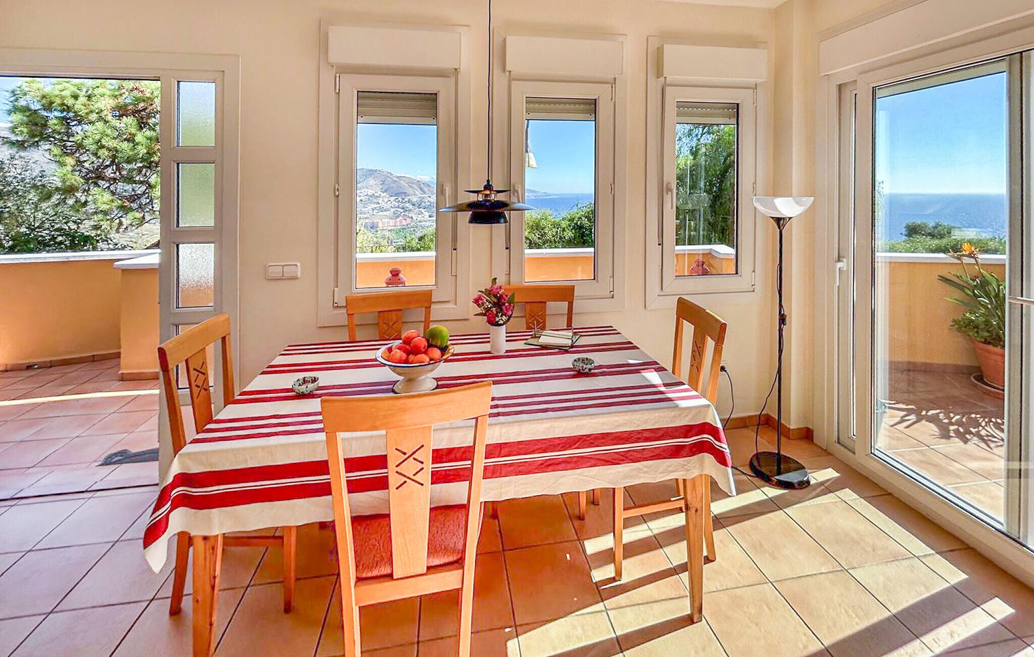 Un coin repas avec une table dressée pour quatre personnes, entourée de chaises, et offrant une vue sur la mer grâce à de grandes fenêtres.