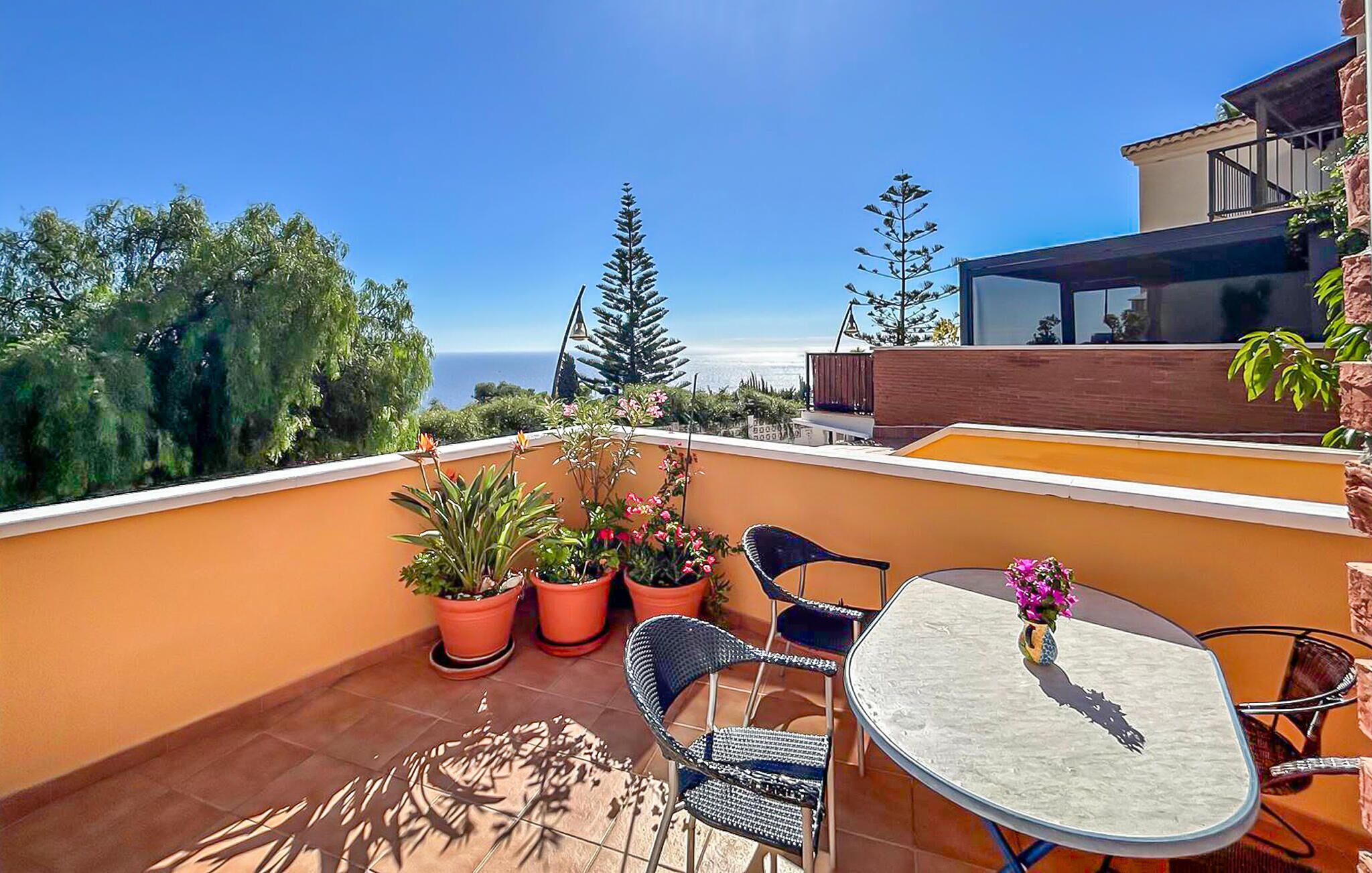 A rooftop terrace with a table and chairs, potted plants, and a view of the ocean.