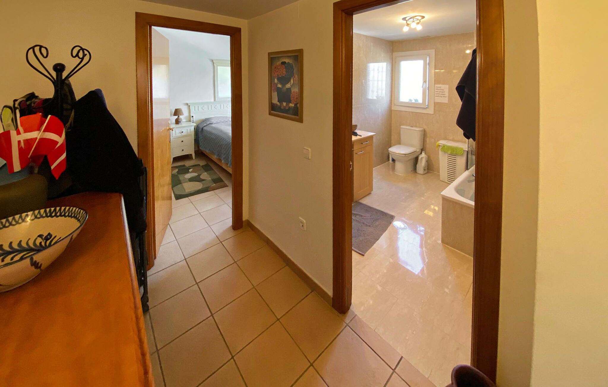 Een gang die leidt naar een slaapkamer met een bed, nachtkastje en een schilderij. Een badkamer met toilet, wastafel en bad is zichtbaar door een geopende deur.