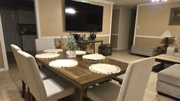 Un comedor con una mesa de madera, sillas blancas y un televisor montado en la pared.