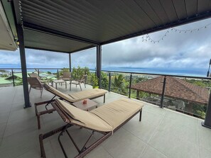 Un espace extérieur aménagé, abrité et doté de chaises longues et d’une table, offrant une vue panoramique sur l’océan et une ville située au loin.