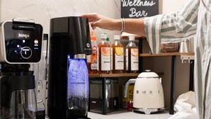 Een keuken met een koffiezetapparaat, een waterdispenser en een koffiemachine. Er staan flessen drank op het aanrecht.