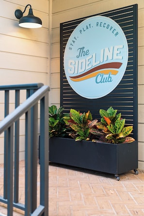 Une enseigne pour «Â
The Sideline Club », avec le slogan «Â
RESTEZ. JOUEZ. REPOSEZ-VOUS. » et un bac à fleurs orné de feuillage coloré.