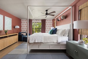 Une chambre Ă coucher avec un lit Ă baldaquin, un ventilateur de plafond, une vue sur la verdure et une table de chevet avec une lampe.