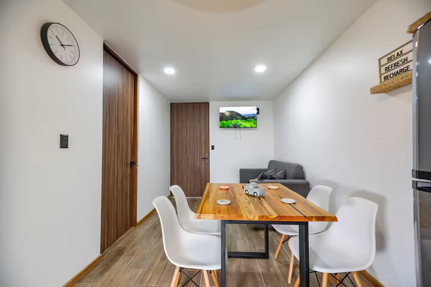 Un comedor moderno con una mesa de madera, sillas blancas, un reloj y un televisor mostrando un paisaje.