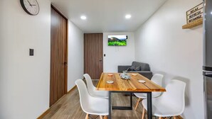 Un comedor moderno con una mesa de madera, sillas blancas, un reloj y un televisor mostrando un paisaje.