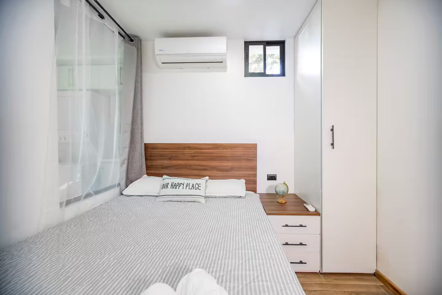 A bedroom with a bed, a nightstand, a wardrobe, an air conditioning unit, and a window with curtains.
