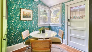 Une pièce avec une table ronde, deux chaises, une plante en pot, un tableau encadré au mur et une fenêtre donnant sur un paysage verdoyant.