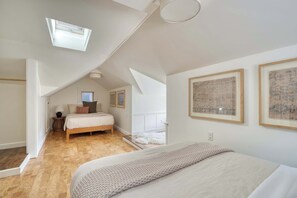 Et soveværelse med skråvæg, ovenlysvindue, seng med hvid dyne, sengebord med lampe og to billeder hængende på væggen.