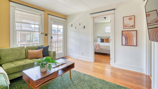 A living room with a green sofa, a wooden coffee table, and a potted plant. There is a sliding door leading to a bedroom with a bed and a nightstand. - 3BR/2BA • Patio • Steps to Mississippi Ave (Portland)