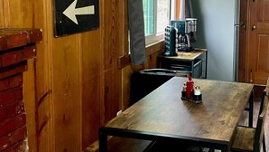 A rustic cabin interior with a wooden dining table, a refrigerator, and a coffee maker. A sign indicates the direction to the East and Route 66. - Rustic Route 66 Homestead (Parks)