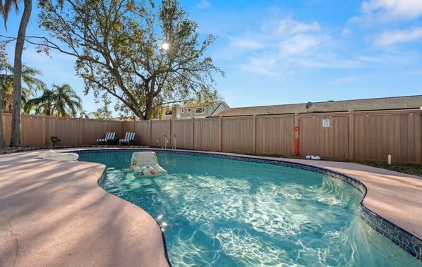 A swimming pool with a wooden fence, lounge chairs, and a tree in the background.